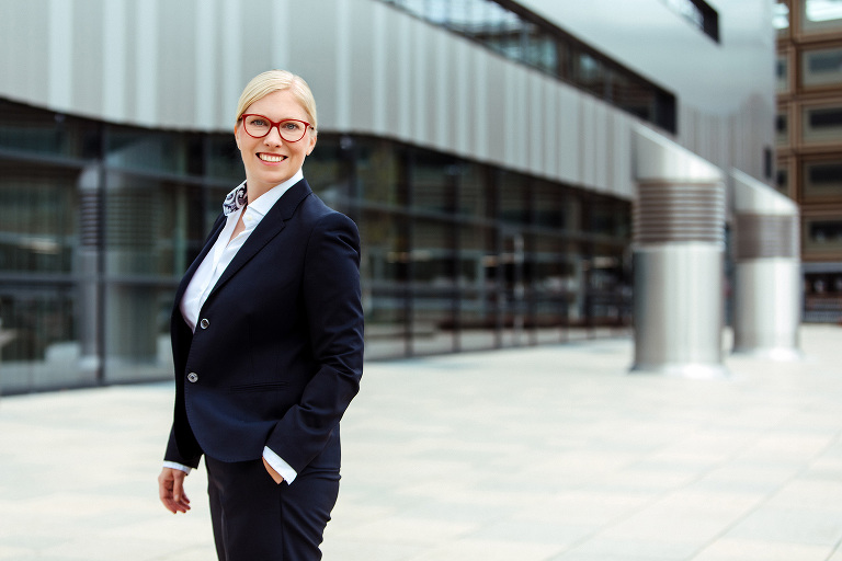 Bewerbungsbild einer Frau. Sie steht vor einem großen Gebäude mit einer Glas-Metallfassade und schaut lächelnd auf den Betrachter. Sie trägt eine weiße Bluse zu einem dunkelblauen Hosenanzug und eine rote Brille.